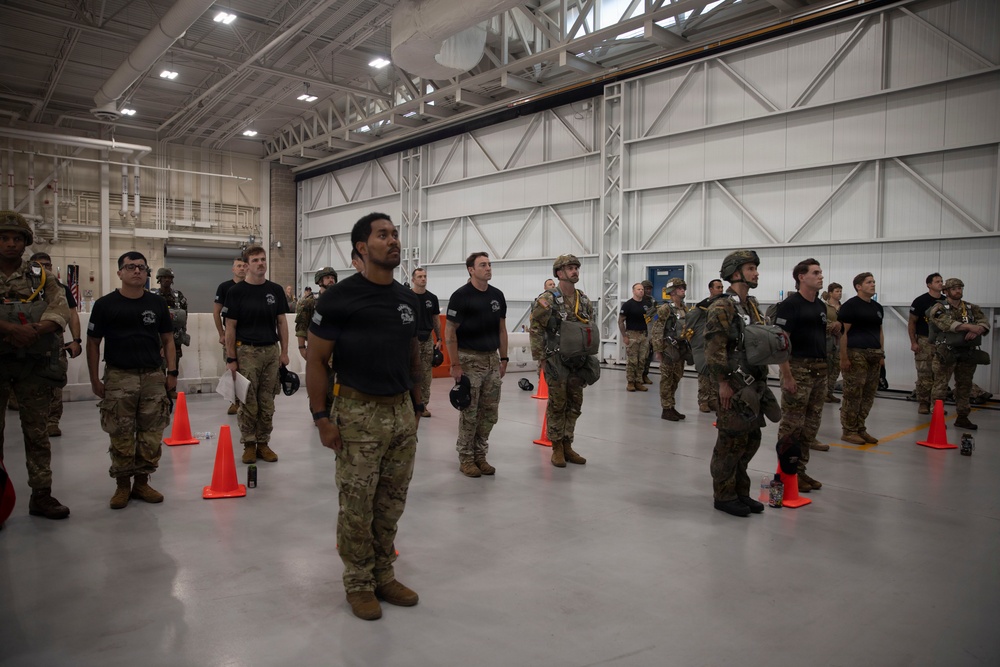 A group of military personnel are assembled in formation inside a large aircraft hangar. The personnel are wearing a mix of black unit t-shirts and full MultiCam/OCP combat uniforms, with some individuals equipped with parachute rigs and tactical gear. Orange traffic cones are used to mark spacing positions on the hangar floor, suggesting a formal muster, briefing, or pre-jump inspection.