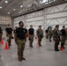 A group of military personnel are assembled in formation inside a large aircraft hangar. The personnel are wearing a mix of black unit t-shirts and full MultiCam/OCP combat uniforms, with some individuals equipped with parachute rigs and tactical gear. Orange traffic cones are used to mark spacing positions on the hangar floor, suggesting a formal muster, briefing, or pre-jump inspection.