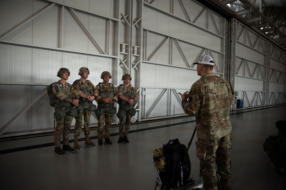 A military instructor in multicam uniform and white cap addresses four soldiers equipped with parachute gear inside a large aircraft hangar. The four soldiers are wearing helmets and parachute harnesses, standing in a line while receiving what appears to be a pre-jump briefing. The setting is a modern hangar with large metal doors and structural cross-bracing visible in the background.