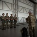 A military instructor in multicam uniform and white cap addresses four soldiers equipped with parachute gear inside a large aircraft hangar. The four soldiers are wearing helmets and parachute harnesses, standing in a line while receiving what appears to be a pre-jump briefing. The setting is a modern hangar with large metal doors and structural cross-bracing visible in the background.