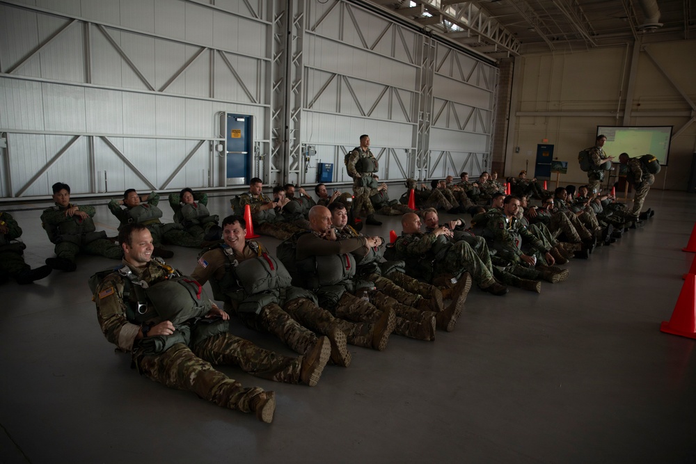 A large group of military personnel in combat uniforms and flight suits are seated on the floor of an aircraft hangar, appearing to receive a briefing or pre-mission instruction. They are equipped with parachute harnesses and tactical gear, suggesting a airborne or jump operation. One standing individual appears to be addressing the group near orange traffic cones used as markers.