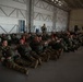 A large group of military personnel in combat uniforms and flight suits are seated on the floor of an aircraft hangar, appearing to receive a briefing or pre-mission instruction. They are equipped with parachute harnesses and tactical gear, suggesting a airborne or jump operation. One standing individual appears to be addressing the group near orange traffic cones used as markers.