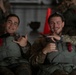 Two young military personnel in camouflage uniforms and parachute harnesses pose and smile for the camera in what appears to be a hangar or staging area. Both are wearing multicam/OCP pattern uniforms with gray parachute rigs featuring red release handles. They appear relaxed and in good spirits, likely preparing for or having completed a airborne/parachute operation.