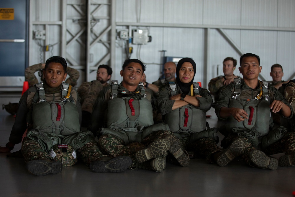 A group of military personnel in parachute/jump gear are seated on the floor of what appears to be a hangar facility. Four individuals are prominently visible in the foreground wearing olive-drab jump harnesses with red release handles over camouflage uniforms, including one female wearing a hijab. Additional personnel in different uniforms are visible in the background.