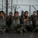 A group of military personnel in parachute/jump gear are seated on the floor of what appears to be a hangar facility. Four individuals are prominently visible in the foreground wearing olive-drab jump harnesses with red release handles over camouflage uniforms, including one female wearing a hijab. Additional personnel in different uniforms are visible in the background.