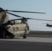 The image shows two U.S. Army CH-47 Chinook tandem-rotor helicopters on what appears to be a military airfield or tarmac. The foreground Chinook is on the ground with rotors spinning and at least one crew member visible near the rear ramp, while a second Chinook is airborne in the background. The lighting suggests early morning or late afternoon conditions.