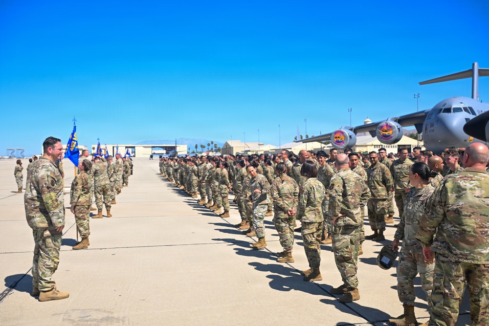 A large military formation is assembled on an airfield tarmac, with personnel wearing OCP (Operational Camouflage Pattern) uniforms. Blue unit guidon flags are visible on the left side, and a military transport aircraft (appears to be a C-17 Globemaster III) is visible in the background. The event appears to be a formal military ceremony, possibly a deployment sendoff, homecoming, or unit formation.