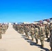 A large military formation is assembled on an airfield tarmac, with personnel wearing OCP (Operational Camouflage Pattern) uniforms. Blue unit guidon flags are visible on the left side, and a military transport aircraft (appears to be a C-17 Globemaster III) is visible in the background. The event appears to be a formal military ceremony, possibly a deployment sendoff, homecoming, or unit formation.