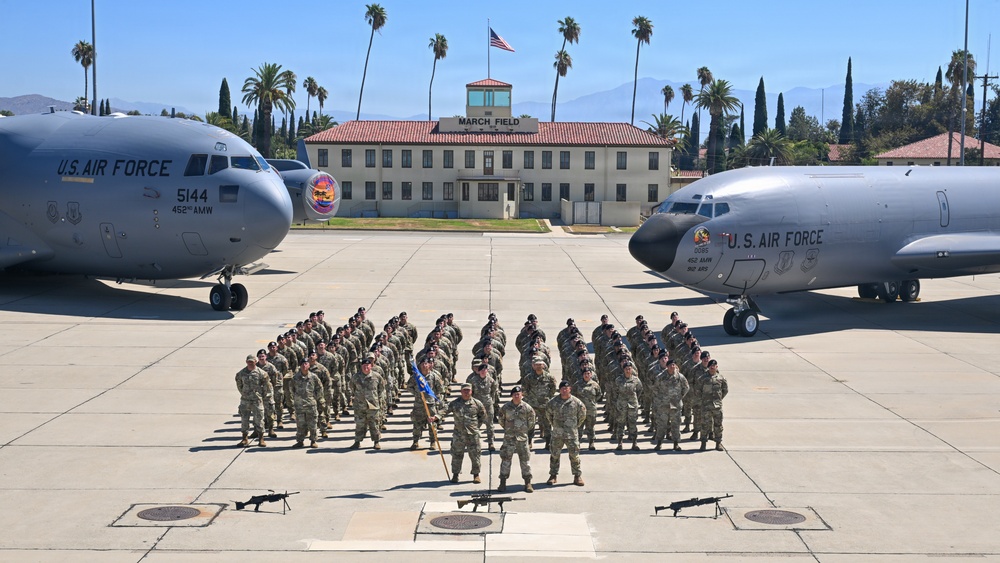 A formal military unit photograph taken on the flight line at March Field Air Reserve Base in California. A large formation of U.S. Air Force personnel in camouflage uniforms are posed between two aircraft — a C-17 Globemaster III (tail number 5144, 452nd AMW) on the left and a KC-135 Stratotanker on the right. The historic March Field building with its red-tile roof and American flag is visible in the background.