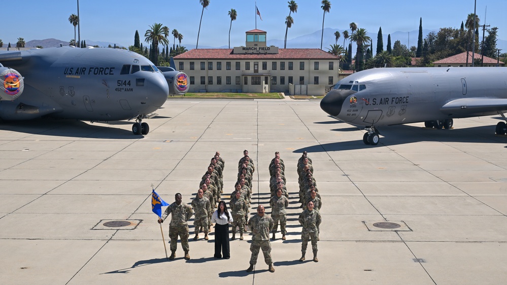 A group of U.S. Air Force personnel pose in formation on the flight line at March Field Air Reserve Base in California. Two large U.S. Air Force aircraft flank the group on either side, with the historic March Field building visible in the background. A civilian woman in white and black attire stands at the front center of the formation alongside uniformed airmen, one of whom holds a unit guidon flag.