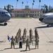 A group of U.S. Air Force personnel pose in formation on the flight line at March Field Air Reserve Base in California. Two large U.S. Air Force aircraft flank the group on either side, with the historic March Field building visible in the background. A civilian woman in white and black attire stands at the front center of the formation alongside uniformed airmen, one of whom holds a unit guidon flag.