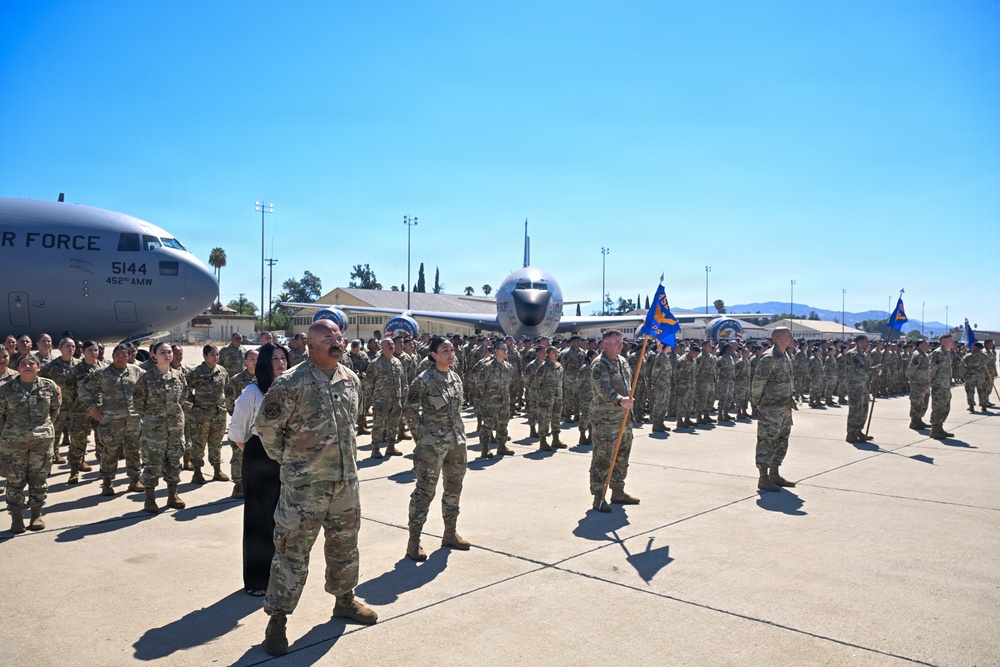 A large formation of U.S. Air Force personnel in OCP (Operational Camouflage Pattern) uniforms are assembled on a flight line apron during what appears to be a formal military ceremony. A C-17 Globemaster III aircraft marked '5144' and '452nd AMW' is visible in the background, indicating this is likely March Air Reserve Base in California. Multiple unit guidon flags in blue are visible, and a civilian woman stands alongside a senior military member in the foreground.