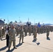 A large formation of U.S. Air Force personnel in OCP (Operational Camouflage Pattern) uniforms are assembled on a flight line apron during what appears to be a formal military ceremony. A C-17 Globemaster III aircraft marked '5144' and '452nd AMW' is visible in the background, indicating this is likely March Air Reserve Base in California. Multiple unit guidon flags in blue are visible, and a civilian woman stands alongside a senior military member in the foreground.