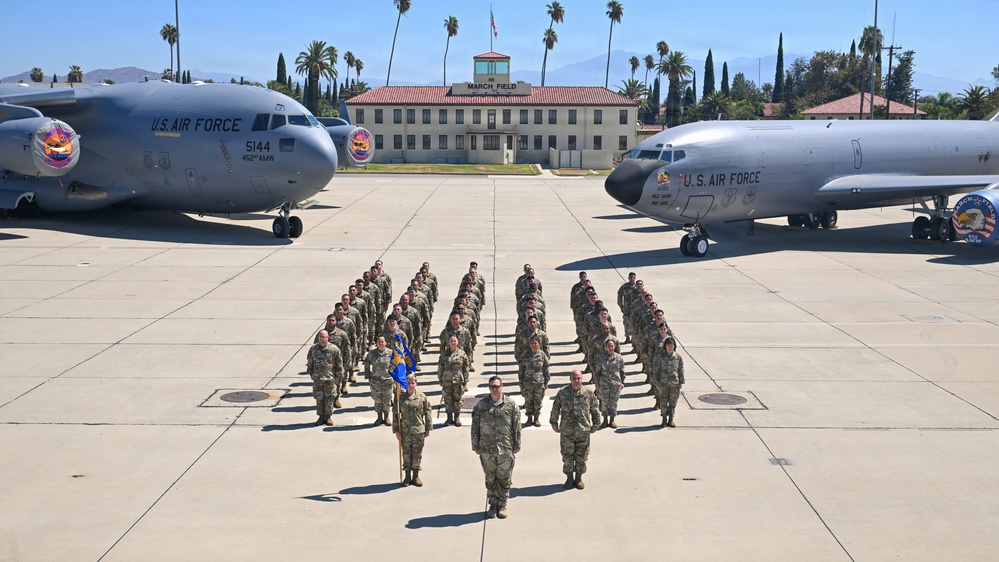 A formal military formation photograph taken on the flight line at March Field, showing U.S. Air Force personnel arranged in a V-shaped formation on the tarmac. Two U.S. Air Force aircraft flank the formation — a C-17 Globemaster III on the left (tail number 5144, 452nd AMW) and a KC-135 Stratotanker on the right. The historic March Field administration building is visible in the background, surrounded by palm trees.