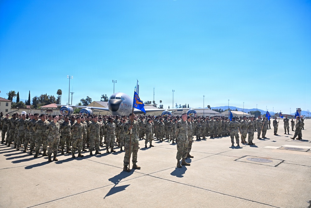 A large military formation is assembled on an airfield tarmac, with hundreds of personnel in camouflage uniforms standing in organized rows. Blue unit guidon flags are visible among the formation, suggesting a formal military ceremony or parade. A large military aircraft is parked in the background, with base infrastructure including hangars and light towers visible.
