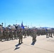 A large military formation is assembled on an airfield tarmac, with hundreds of personnel in camouflage uniforms standing in organized rows. Blue unit guidon flags are visible among the formation, suggesting a formal military ceremony or parade. A large military aircraft is parked in the background, with base infrastructure including hangars and light towers visible.
