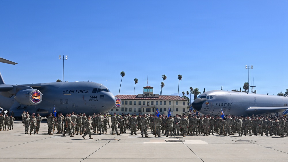 A large U.S. Air Force military formation ceremony is taking place on the flight line at March Field (March Air Reserve Base) in California. Hundreds of uniformed personnel are assembled in formation on the tarmac between two large military aircraft. The historic March Field headquarters building is visible in the background, flanked by palm trees.