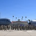 A large U.S. Air Force military formation ceremony is taking place on the flight line at March Field (March Air Reserve Base) in California. Hundreds of uniformed personnel are assembled in formation on the tarmac between two large military aircraft. The historic March Field headquarters building is visible in the background, flanked by palm trees.