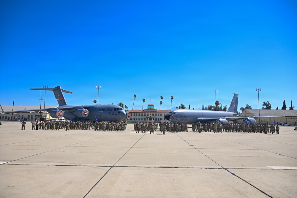 A large military formation ceremony is taking place on an airfield tarmac, with hundreds of uniformed personnel assembled in organized groups. Two U.S. Air Force aircraft are prominently displayed behind the formation — a C-17 Globemaster III on the left and what appears to be a KC-135 Stratotanker on the right. The setting includes palm trees, base buildings, and light poles consistent with a Southern California military installation.