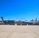 A large military formation ceremony is taking place on an airfield tarmac, with hundreds of uniformed personnel assembled in organized groups. Two U.S. Air Force aircraft are prominently displayed behind the formation — a C-17 Globemaster III on the left and what appears to be a KC-135 Stratotanker on the right. The setting includes palm trees, base buildings, and light poles consistent with a Southern California military installation.