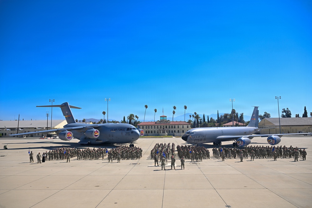 A large U.S. Air Force formation ceremony is taking place on a flight line apron, with hundreds of uniformed military personnel assembled between two aircraft. The scene features a C-17 Globemaster III on the left and a KC-135 Stratotanker on the right, both bearing unit insignia on their engines. The setting appears to be a Southern California air base, identifiable by the palm trees, Spanish Colonial Revival architecture, and mountain backdrop.