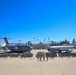 A large U.S. Air Force formation ceremony is taking place on a flight line apron, with hundreds of uniformed military personnel assembled between two aircraft. The scene features a C-17 Globemaster III on the left and a KC-135 Stratotanker on the right, both bearing unit insignia on their engines. The setting appears to be a Southern California air base, identifiable by the palm trees, Spanish Colonial Revival architecture, and mountain backdrop.