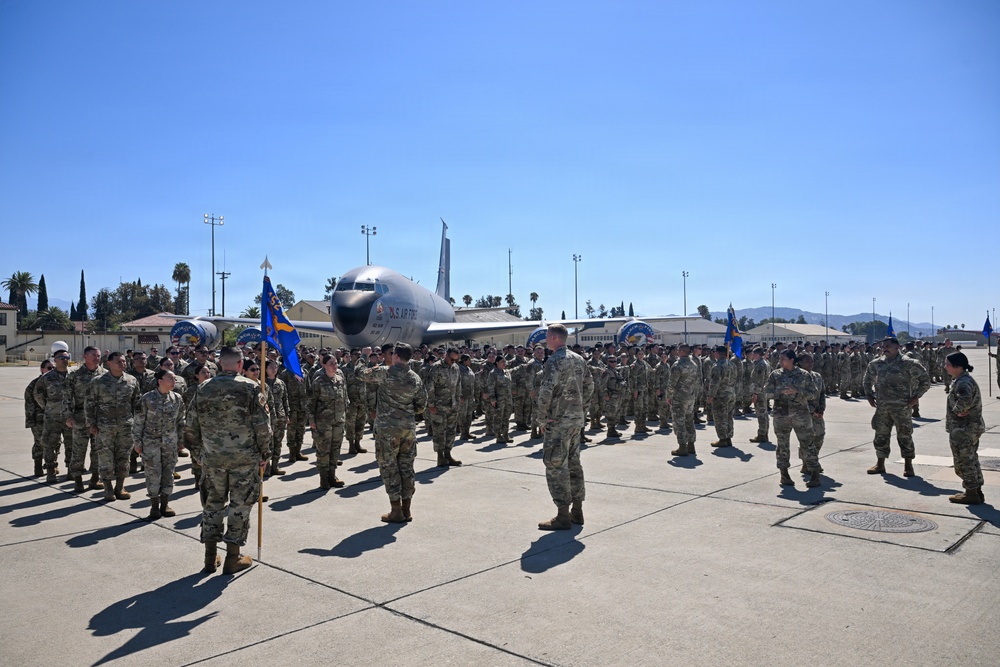 A large formation of U.S. Air Force personnel in OCP (Operational Camouflage Pattern) uniforms are assembled on a flight line apron during what appears to be a formal military ceremony or parade. A U.S. Air Force aircraft, identifiable as a KC-135 or similar large jet, is parked in the background. The setting appears to be a U.S. Air Force base, likely in Southern California based on the palm trees, mountains, and architectural style.