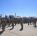 A large formation of U.S. Air Force personnel in OCP (Operational Camouflage Pattern) uniforms are assembled on a flight line apron during what appears to be a formal military ceremony or parade. A U.S. Air Force aircraft, identifiable as a KC-135 or similar large jet, is parked in the background. The setting appears to be a U.S. Air Force base, likely in Southern California based on the palm trees, mountains, and architectural style.