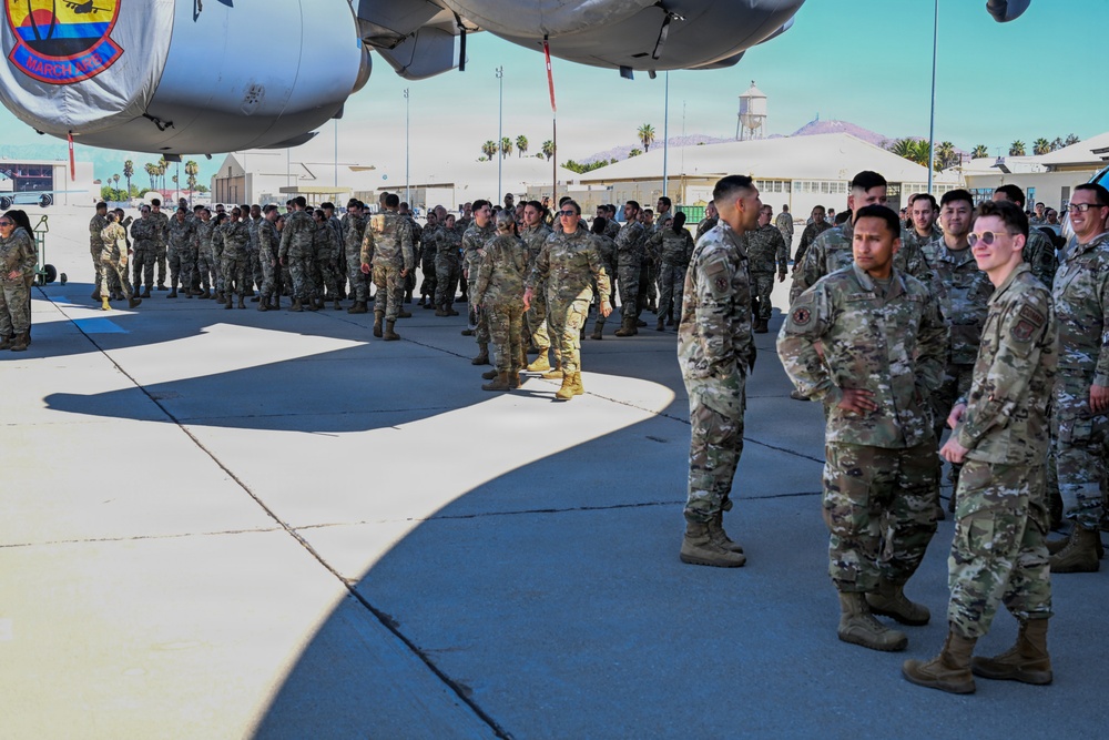 A large formation of military personnel in OCP (Operational Camouflage Pattern) uniforms are gathered on an aircraft flight line apron, standing beneath the fuselage of a large military transport aircraft. A unit patch is visible on the aircraft, and the setting appears to be a U.S. Air Force Reserve or Air National Guard base with hangars, palm trees, a water tower, and mountains visible in the background. The gathering appears to be a formal unit assembly, ceremony, or briefing event.