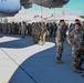 A large formation of military personnel in OCP (Operational Camouflage Pattern) uniforms are gathered on an aircraft flight line apron, standing beneath the fuselage of a large military transport aircraft. A unit patch is visible on the aircraft, and the setting appears to be a U.S. Air Force Reserve or Air National Guard base with hangars, palm trees, a water tower, and mountains visible in the background. The gathering appears to be a formal unit assembly, ceremony, or briefing event.