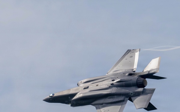 A fifth-generation stealth fighter jet, identifiable as an F-35 Lightning II, is captured in flight performing a banking maneuver against a partly cloudy sky. The aircraft is photographed from below and slightly behind, revealing its distinctive delta-wing planform and stealth-optimized airframe. Vapor trails are visible streaming from the wingtips, indicating high-speed or high-G flight conditions.