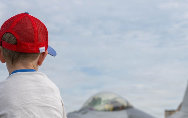 A young boy wearing a red baseball cap and white t-shirt is viewed from behind as he gazes at a gray military fighter jet on an airfield tarmac. The scene appears to be an air show or military base open house event. The composition captures a classic moment of childhood wonder toward aviation and military aircraft.
