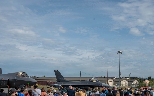 A large crowd of civilians gathers at a military air show on a flight line, viewing two parked fighter jets up close. The aircraft visible include what appears to be an F-35 Lightning II on the left and an F-16 Fighting Falcon on the right, both in gray military livery. The event appears to be an open house or public airshow at a military installation, likely in Alaska based on the terrain and vegetation.