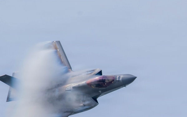 A modern stealth fighter jet, consistent with an F-35 Lightning II, is captured in high-speed flight at a banked angle. A prominent vapor cone or condensation cloud is visible wrapping around the fuselage, indicating high-speed, low-altitude flight through humid air. The aircraft appears to be performing an aerobatic or demonstration maneuver.