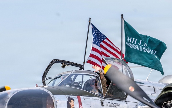 A restored World War II-era B-25 Mitchell bomber named 'Million Airess' is displayed on a tarmac, featuring classic nose art of a pin-up figure. The aircraft bears the 'Million Air' branding on its fuselage and is flying both an American flag and a green 'Million Air' company flag. A person is visible waving from the cockpit window.