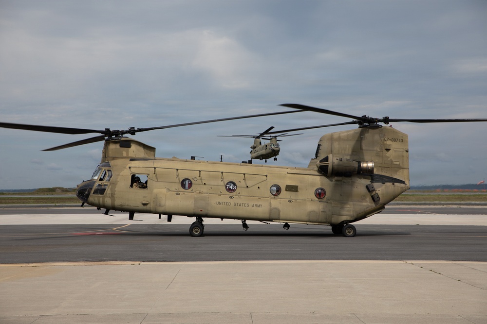 A United States Army CH-47 Chinook helicopter is parked on a tarmac with its rotors spinning, bearing the serial number 07-08743 and side number 743. A second Chinook helicopter is visible in the background, appearing to be in flight or hovering. Both aircraft are painted in standard Army tan/olive drab livery.