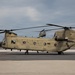 A United States Army CH-47 Chinook helicopter is parked on a tarmac with its rotors spinning, bearing the serial number 07-08743 and side number 743. A second Chinook helicopter is visible in the background, appearing to be in flight or hovering. Both aircraft are painted in standard Army tan/olive drab livery.
