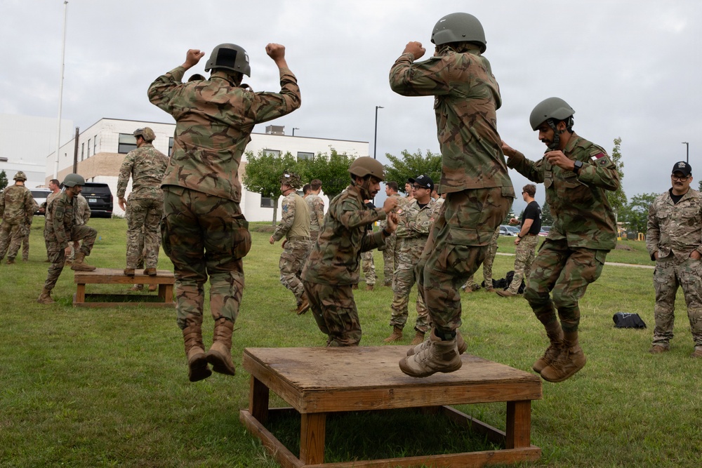 Military personnel in camouflage uniforms and helmets are engaged in a physical training exercise on a grass field near a building. Several soldiers are jumping on and off a wooden platform box, while others observe or participate in similar drills in the background. The activity appears to be a structured combat fitness or obstacle course training event.