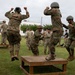 Military personnel in camouflage uniforms and helmets are engaged in a physical training exercise on a grass field near a building. Several soldiers are jumping on and off a wooden platform box, while others observe or participate in similar drills in the background. The activity appears to be a structured combat fitness or obstacle course training event.