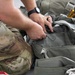 A person wearing military camouflage (MultiCam/OCP pattern) trousers is handling what appears to be a parachute harness or tactical gear assembly. The individual is inspecting or adjusting gray nylon straps, buckles, and webbing components laid out on a flat surface. The close-up perspective focuses on the hands and equipment, suggesting a gear inspection or packing procedure.