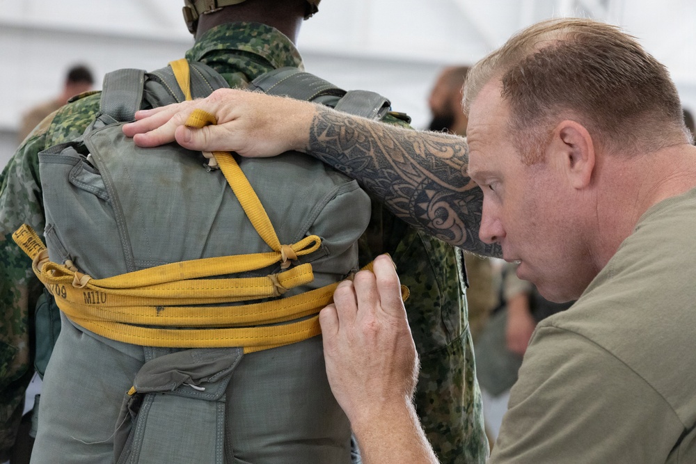 A military instructor or rigger with visible tribal tattoos on his forearm is closely inspecting or adjusting a parachute pack on another soldier's back. The soldier wearing the parachute is dressed in a camouflage uniform, and the pack is secured with yellow/gold straps marked with identifiers. This appears to be a pre-jump equipment check or parachute rigging inspection in a training or operational environment.