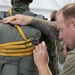A military instructor or rigger with visible tribal tattoos on his forearm is closely inspecting or adjusting a parachute pack on another soldier's back. The soldier wearing the parachute is dressed in a camouflage uniform, and the pack is secured with yellow/gold straps marked with identifiers. This appears to be a pre-jump equipment check or parachute rigging inspection in a training or operational environment.