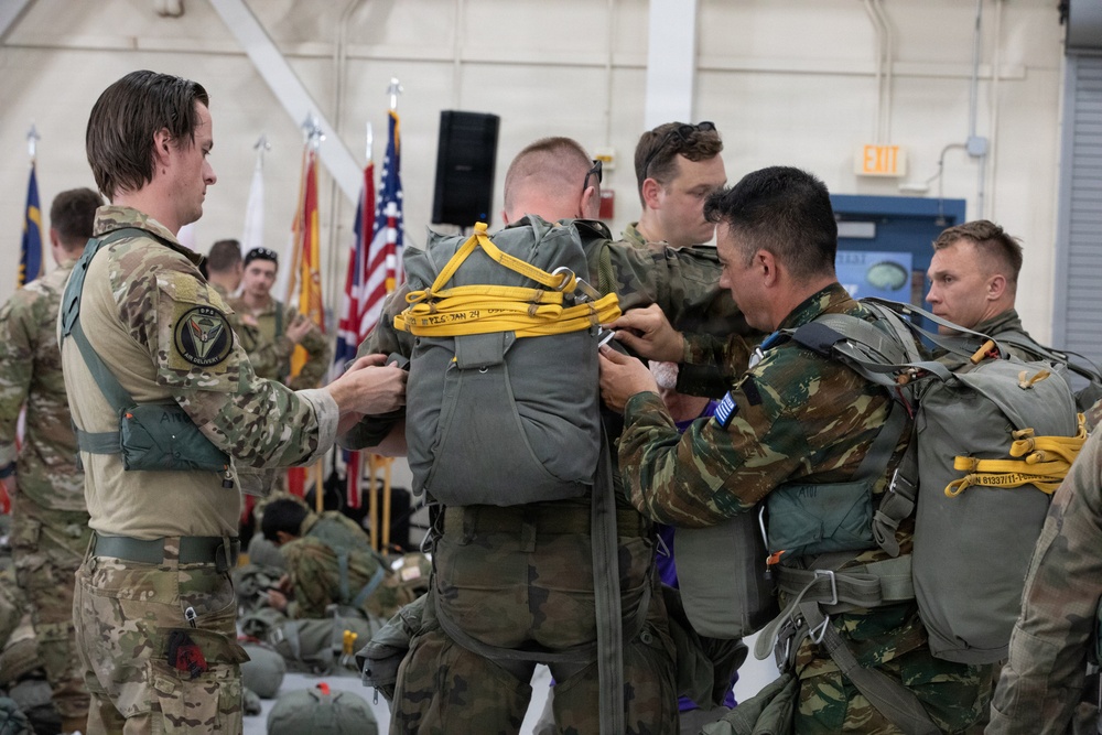 Military personnel in a hangar facility are conducting pre-jump equipment checks, inspecting and adjusting parachute harnesses and rigs on each other. The group includes soldiers in different camouflage patterns, suggesting a multinational or joint training exercise. An American flag and other military unit flags are visible in the background.