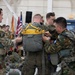 Military personnel in a hangar facility are conducting pre-jump equipment checks, inspecting and adjusting parachute harnesses and rigs on each other. The group includes soldiers in different camouflage patterns, suggesting a multinational or joint training exercise. An American flag and other military unit flags are visible in the background.