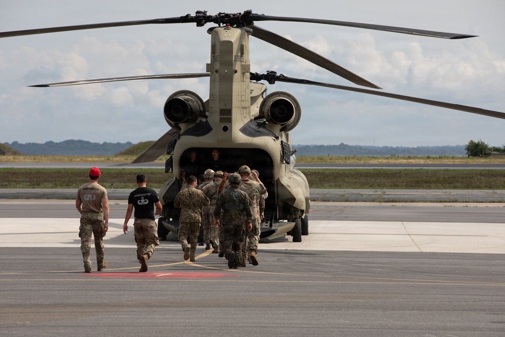 A group of military personnel in combat uniforms are boarding or loading a CH-47 Chinook tandem-rotor helicopter on a paved airfield tarmac. The soldiers are approaching the rear ramp of the aircraft, with some already inside. The setting appears to be a military airfield during daytime with partly cloudy skies.