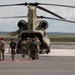 A group of military personnel in combat uniforms are boarding or loading a CH-47 Chinook tandem-rotor helicopter on a paved airfield tarmac. The soldiers are approaching the rear ramp of the aircraft, with some already inside. The setting appears to be a military airfield during daytime with partly cloudy skies.