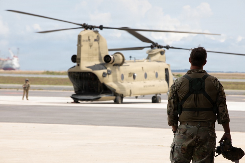 A military personnel in camouflage uniform stands on a tarmac observing a tan/olive-colored CH-47 Chinook tandem-rotor helicopter parked on the flight line. A second uniformed individual is visible in the background near the aircraft. The setting appears to be a military airfield with water and vessels visible in the far background.