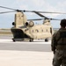 A military personnel in camouflage uniform stands on a tarmac observing a tan/olive-colored CH-47 Chinook tandem-rotor helicopter parked on the flight line. A second uniformed individual is visible in the background near the aircraft. The setting appears to be a military airfield with water and vessels visible in the far background.