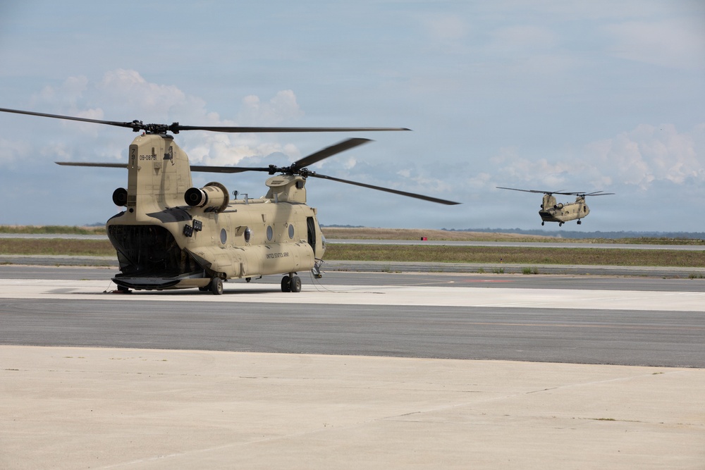 Two U.S. Army CH-47 Chinook tandem-rotor helicopters are visible on what appears to be a military airfield or airport tarmac. The foreground helicopter is parked on the concrete apron with rotors spinning, while a second Chinook is airborne in the background. Both aircraft are painted in desert tan/sand camouflage livery.