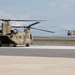 Two U.S. Army CH-47 Chinook tandem-rotor helicopters are visible on what appears to be a military airfield or airport tarmac. The foreground helicopter is parked on the concrete apron with rotors spinning, while a second Chinook is airborne in the background. Both aircraft are painted in desert tan/sand camouflage livery.